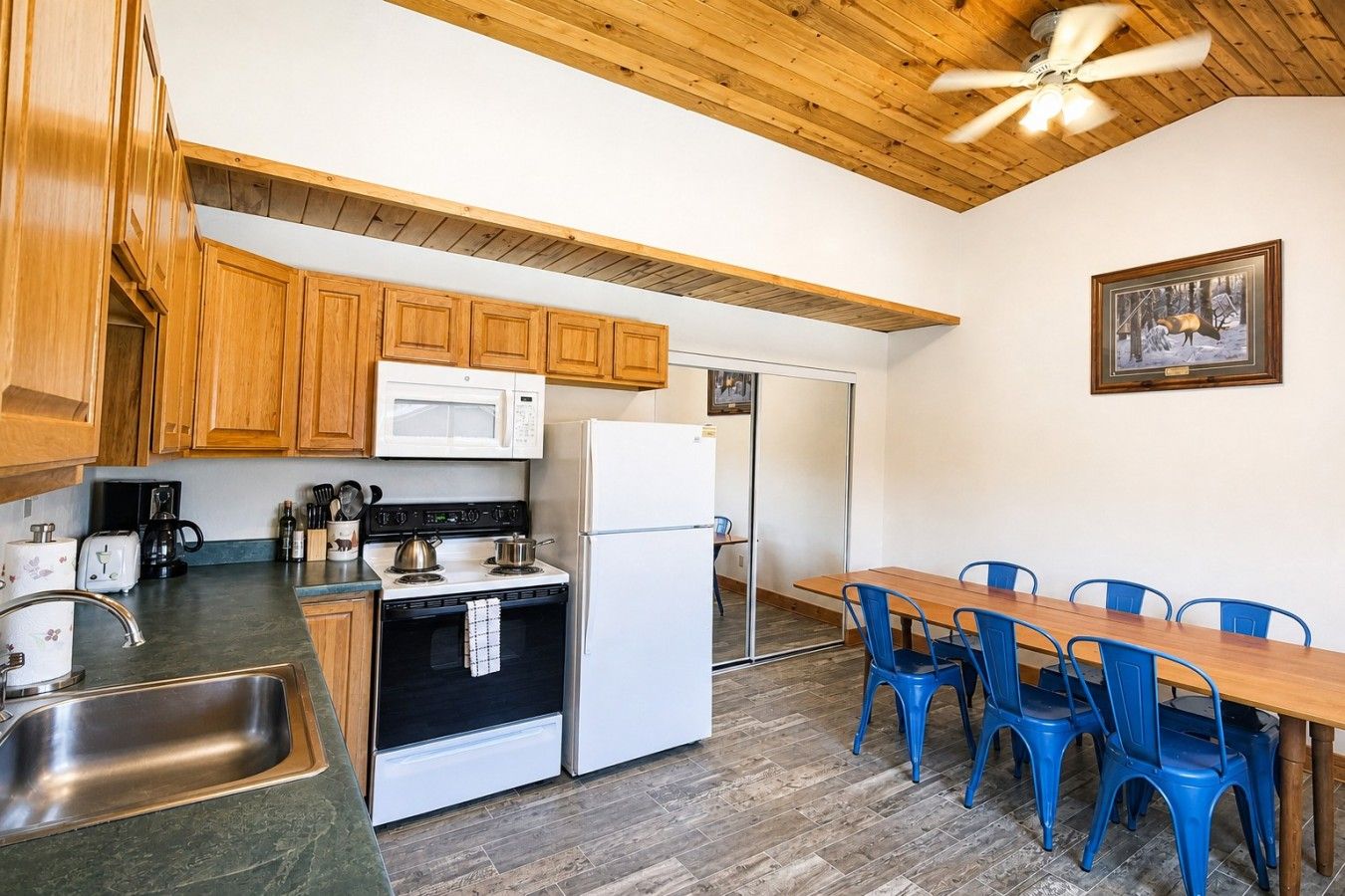 A compact kitchen with wooden cabinets, a microwave, stove, refrigerator, and a dining area with blue chairs.