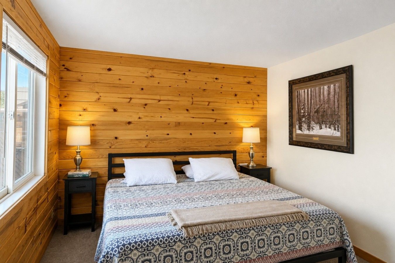 Une chambre avec une tête de lit en bois, une couvre-lit à motifs, deux lampes de chevet, un tableau encadré au mur et une fenêtre sur le côté gauche.