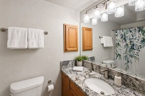 A bathroom with a toilet, a sink with a marble countertop, wooden cabinets, a mirror, and a shower curtain with a floral pattern. - Studio apartment, heart of Grand Lake, Lake views (Grand Lake)