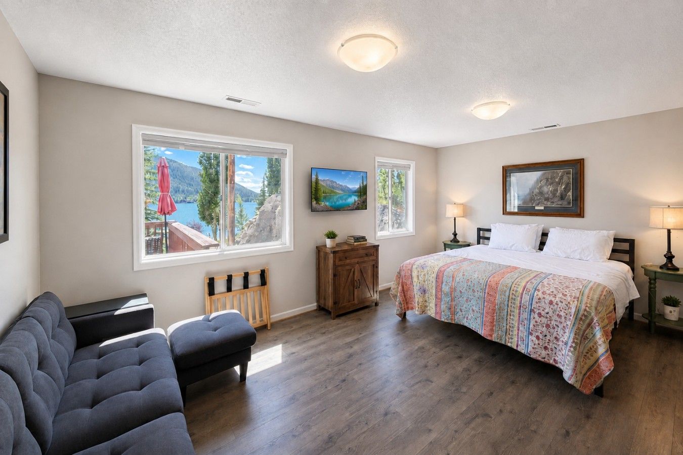 A bedroom with a bed, a sofa, a wooden cabinet, and two windows with views of a lake and mountains.