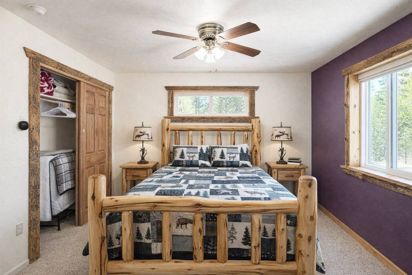 Une chambre avec un lit en bois, deux tables de chevet avec des lampes, un ventilateur de plafond, une fenêtre donnant sur des arbres et un placard avec des vêtements suspendus.