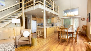 Amplia sala de estar con una escalera de madera, una mesa de comedor con sillas y una zona de cocina.