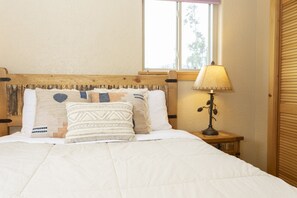 A bed with a white duvet, multiple pillows with various patterns, a wooden headboard, a bedside lamp, and a window with blinds. - Glacial boulder cabin with panoramic mountain view (Grand Lake)