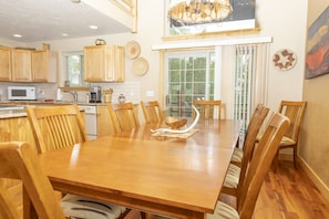 A dining area with a wooden table and chairs, a kitchen in the background, and a large window with curtains. - Glacial boulder cabin with panoramic mountain view (Grand Lake)