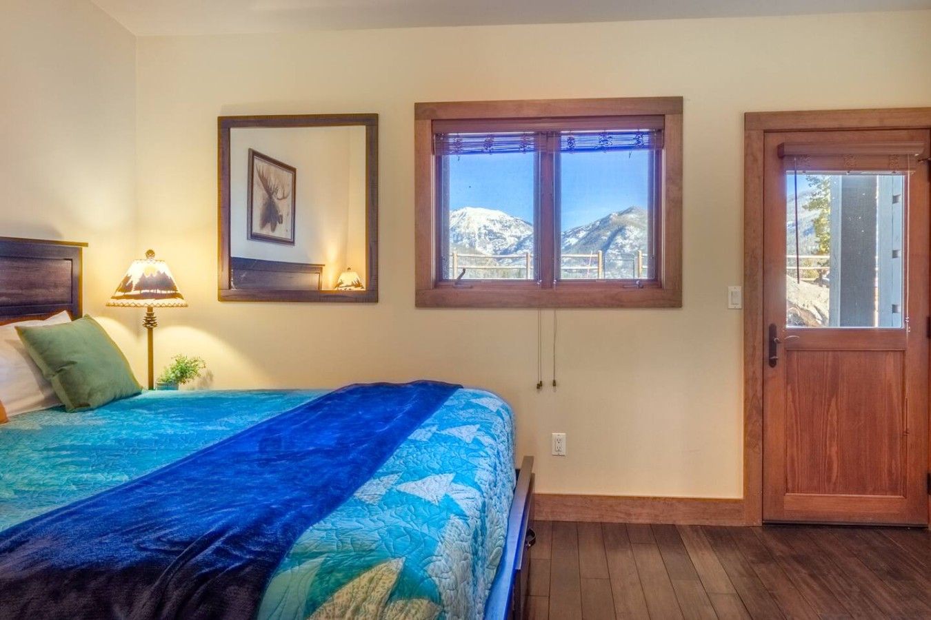 A bedroom with a bed, a window with a mountain view, a wooden door, and a wall-mounted lamp.