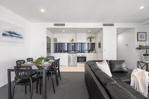 A modern living room with a black leather sofa, a dining area with a table set for four, and a kitchen with white cabinets and blue accents. - Circle on Cavill Ocean View (Surfers Paradise)
