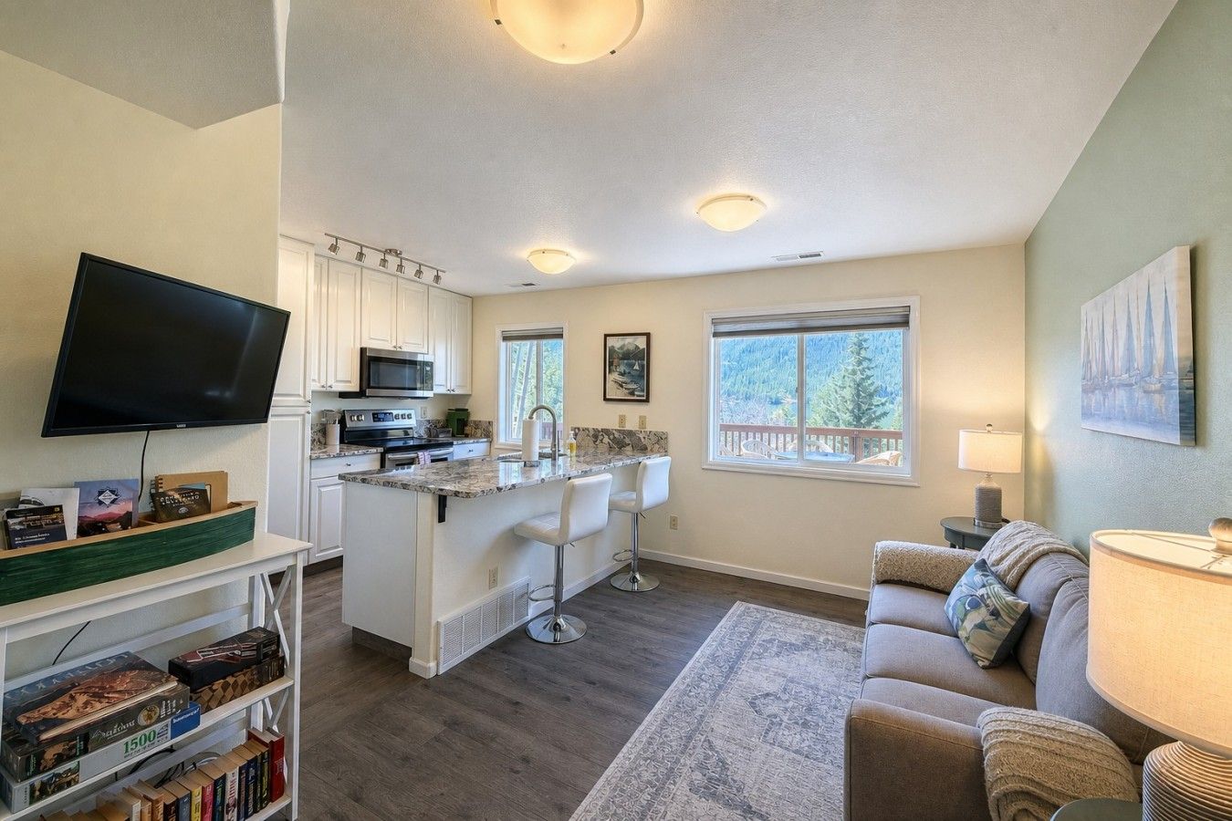 A modern kitchen with white cabinets, a large island with bar stools, a flat-screen TV, and a dining area with a couch and table.