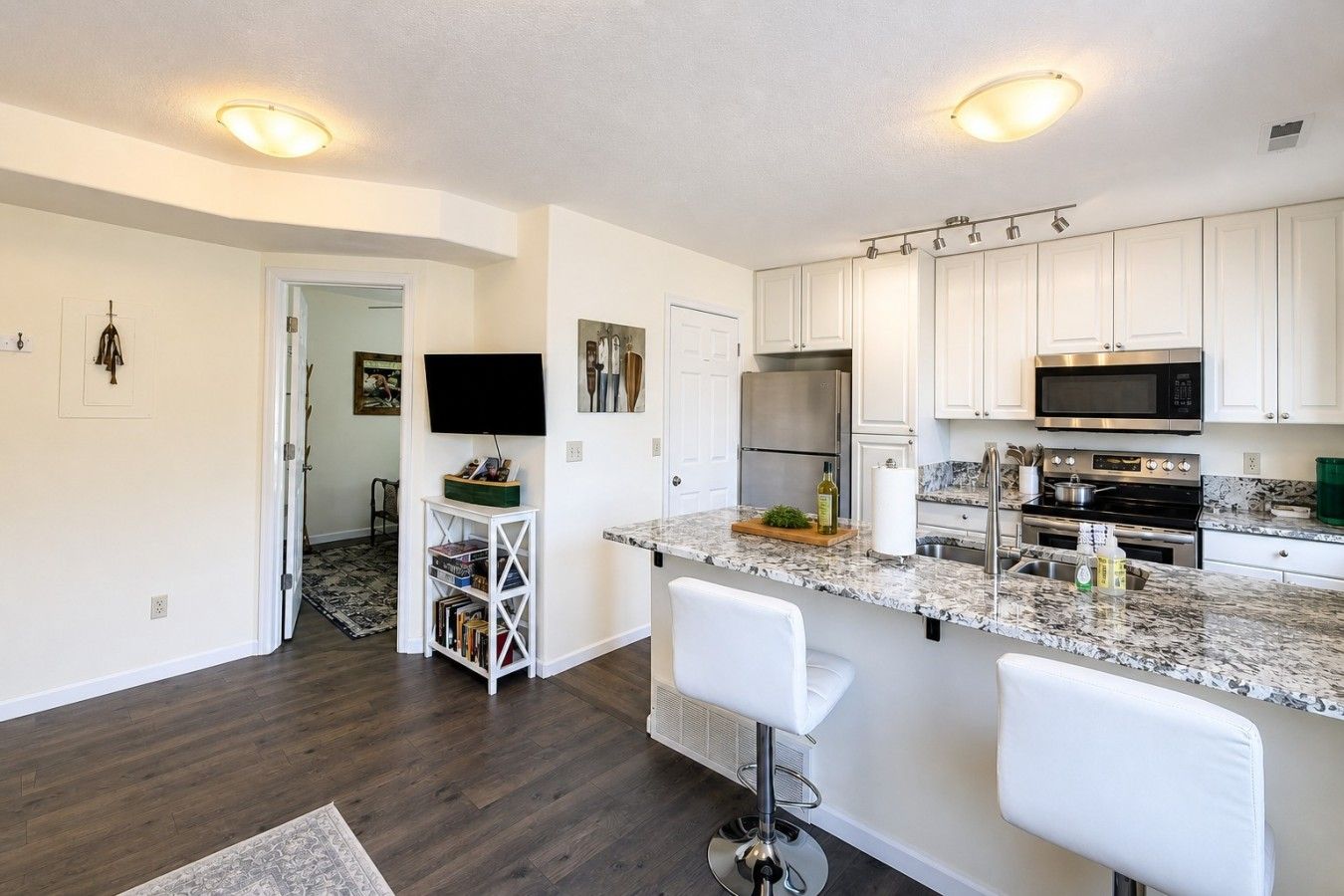 A modern kitchen with white cabinets, a granite countertop, stainless steel appliances, and a bar area with white stools.