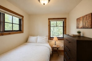 A bedroom with a bed, a dresser, a nightstand with a lamp, and a window with a view of trees. - Lakeside luxury mountain getaway (Grand Lake)