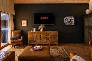Een gezellige woonkamer met een leren fauteuil, een houten kast met lades, een flatscreen-tv waarop 'NETFLIX' staat en een lijst met een schilderij aan de muur.