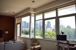 A room with a large window, a sofa, a desk with a computer, and a lamp. - Uptown Condo | Skyline Views + Walkable Third Ward (Charlotte)