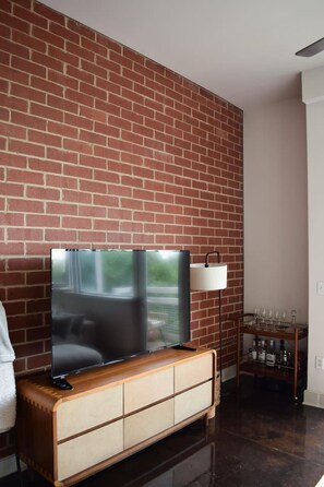 A modern living room with a brick wall, a flat-screen TV on a wooden cabinet, a side table with a lamp, and a shelf with bottles. - Uptown Condo | Skyline Views + Walkable Third Ward (Charlotte)