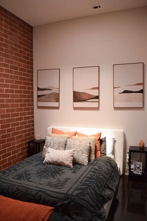 A bedroom with a brick wall, a bed with patterned bedding, and three landscape paintings on the wall. - Uptown Condo | Skyline Views + Walkable Third Ward (Charlotte)