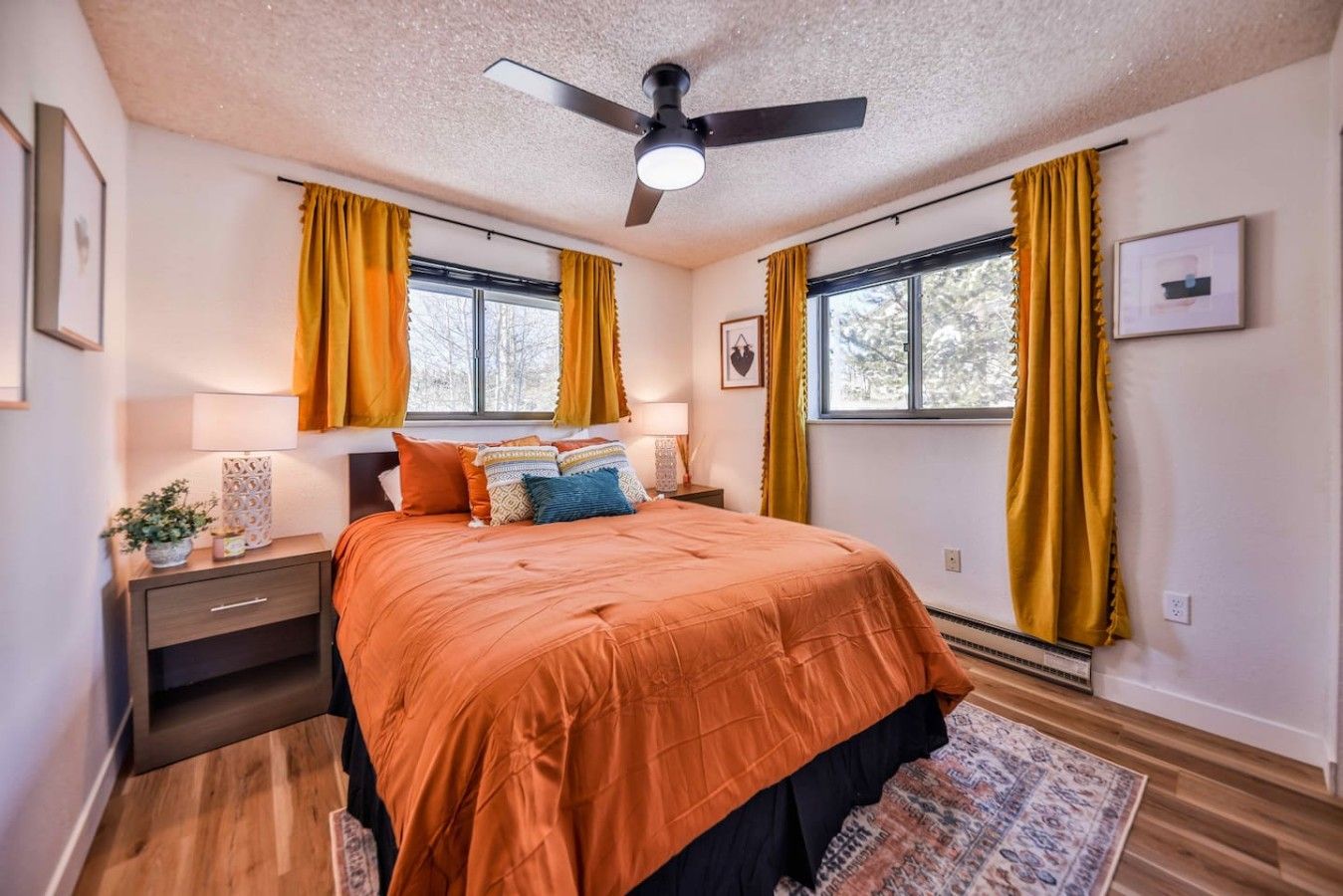 Un dormitorio con cama, mesitas de noche, ventilador de techo y ventanas grandes con cortinas.