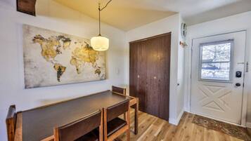 A dining area with a wooden table and chairs, a world map artwork on the wall, a wooden door, and a window with a view.
