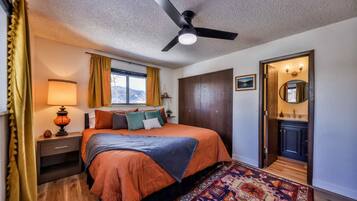 A bedroom with a bed, a nightstand, a lamp, a window with curtains, a ceiling fan, a door leading to a bathroom, and a patterned rug.