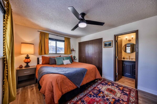 A bedroom with a bed, a nightstand, a lamp, a window with curtains, a ceiling fan, a door leading to a bathroom, and a patterned rug. - Epic Views of Grand Lake | Minutes to RMNP (Grand Lake)