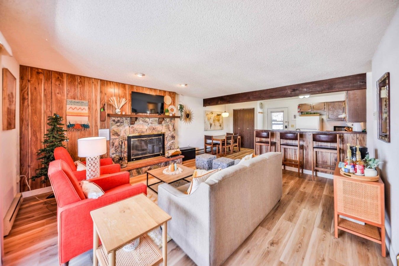 Una sala de estar con chimenea, sofás rojos y grises, una mesa de centro y una zona de cocina con armarios de madera y taburetes de bar.