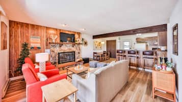 Una sala de estar con chimenea, sofás rojos y grises, una mesa de centro y una zona de cocina con armarios de madera y taburetes de bar.