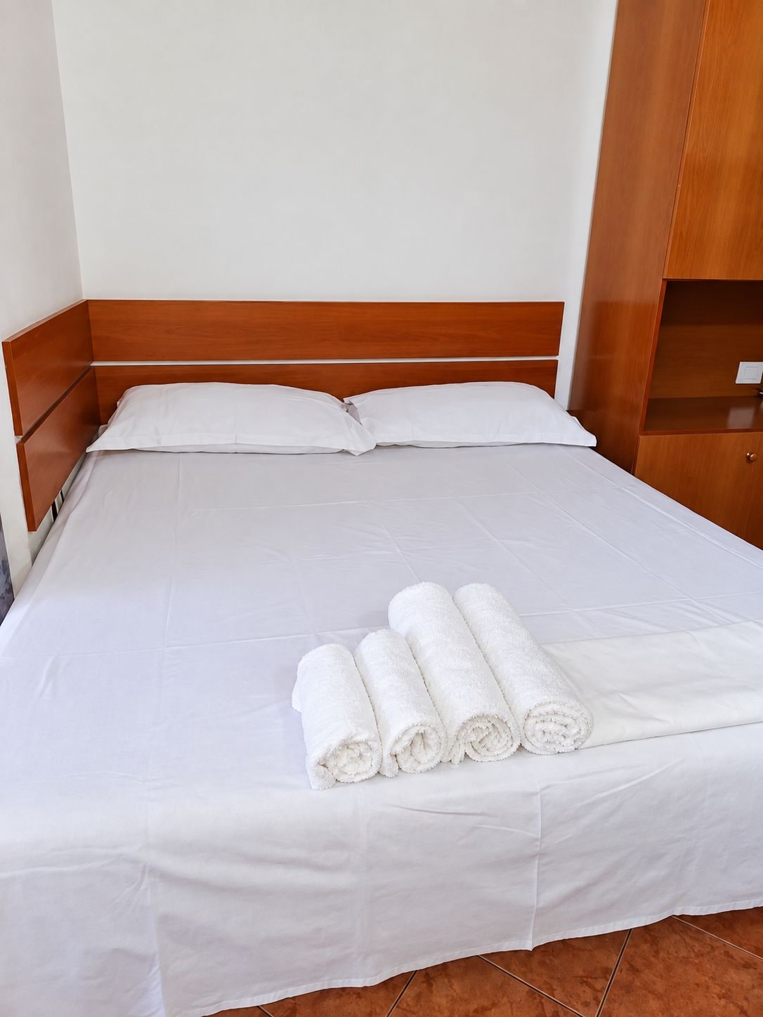 A neatly made bed with white linens and rolled towels, a wooden headboard, and a wooden wardrobe to the right.