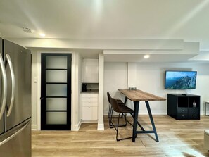 Eine moderne Küche mit Einbaukühlschrank, ein Essbereich mit Holztisch und -stühlen, eine Küchenarbeitsplatte mit Kaffeemaschine und ein an der Wand befestigter Fernseher.