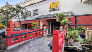 Un restaurante llamado "Aoki Teppanyaki" con fachada roja y blanca, un camino de piedra que conduce a la entrada y un pequeño jardín con plantas y una fuente de piedra.