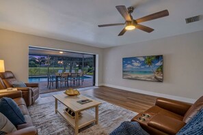 A living room with a ceiling fan, a large window with a view of a pool and outdoor seating, and a painting of a beach scene. - The Pool. The Views. The House. (Sarasota)
