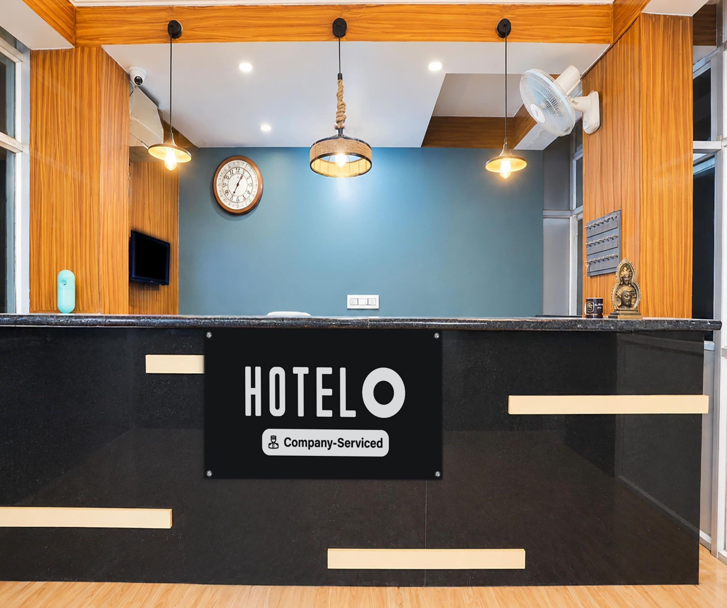 A hotel reception desk with a sign indicating it is company-serviced. There is a clock on the wall, a television, and a decorative item on the counter.