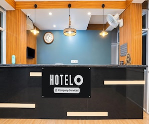 A hotel reception desk with a sign indicating it is company-serviced. There is a clock on the wall, a television, and a decorative item on the counter.