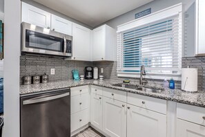 A modern kitchen with white cabinetry, a stainless steel dishwasher, a built-in microwave, and a window with blinds. - 2BR Resort Escape - Pools and Bars (Hilton Head Island)