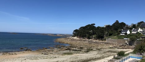 Vista costeira com praia de areia, céu azul límpido e fileira de casas à beira-mar.
