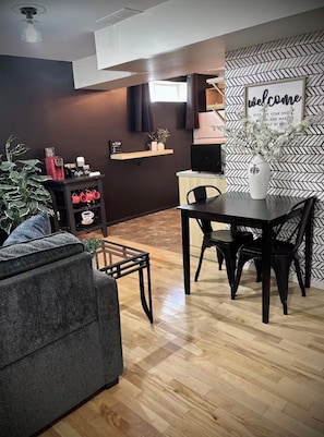 A modern living room with a sofa, a dining table with chairs, a coffee machine, and a wall-mounted TV.