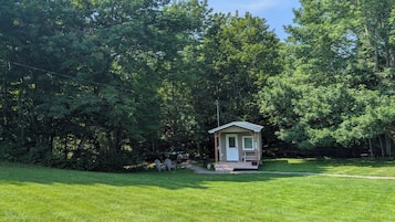 A small wooden cabin with a porch, surrounded by a grassy area and dense trees.