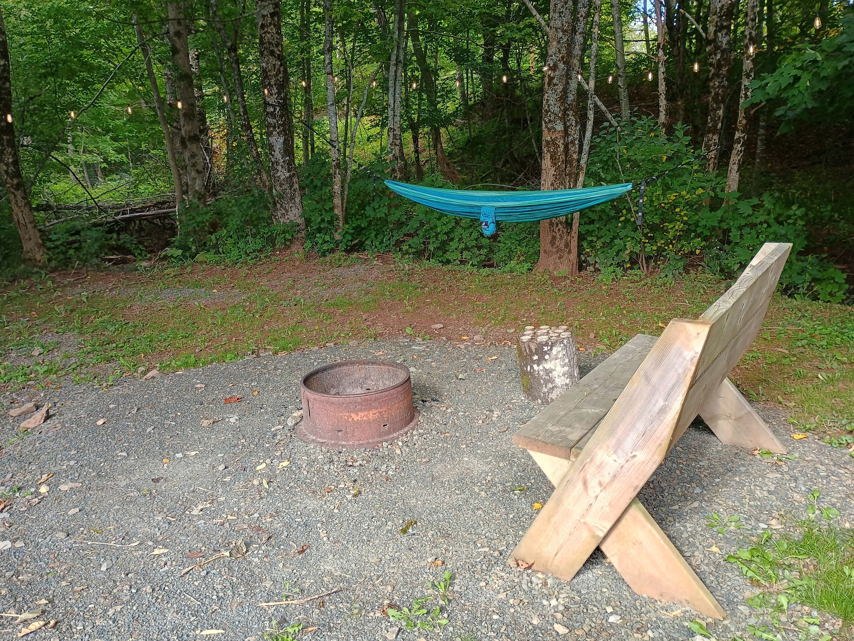 A hammock suspended between trees in a wooded area with a metal fire ring and wooden planks on the ground.
