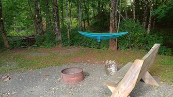 A hammock suspended between trees in a wooded area with a metal fire ring and wooden planks on the ground.