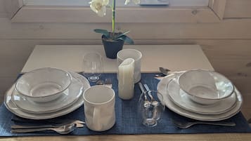 A dining table set with plates, bowls, a mug, and cutlery, with a potted plant and a window in the background.