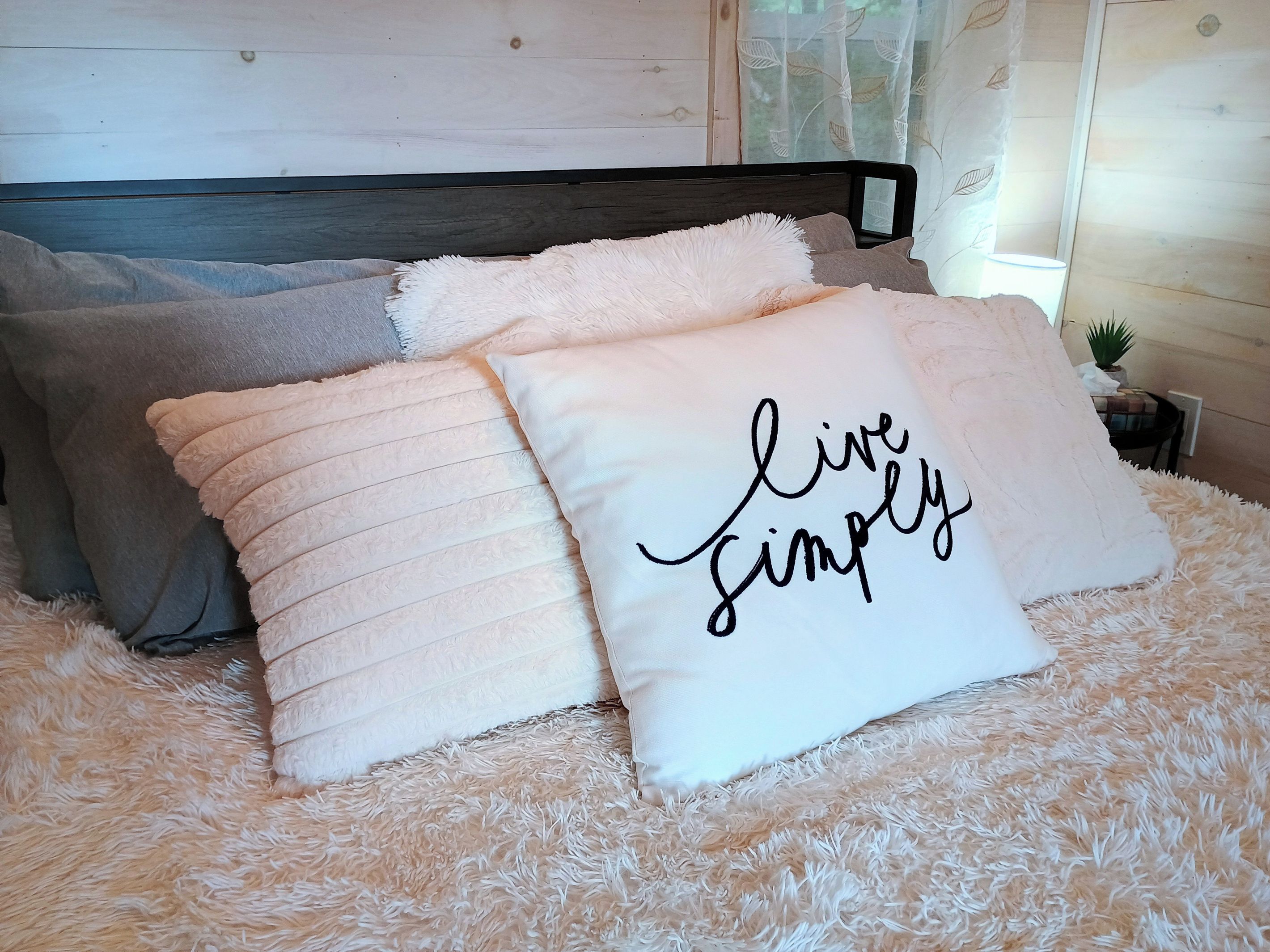A bed with fluffy bedding and pillows, one with the text 'live simply.'