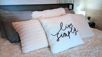A bed with fluffy bedding and pillows, one with the text 'live simply.'