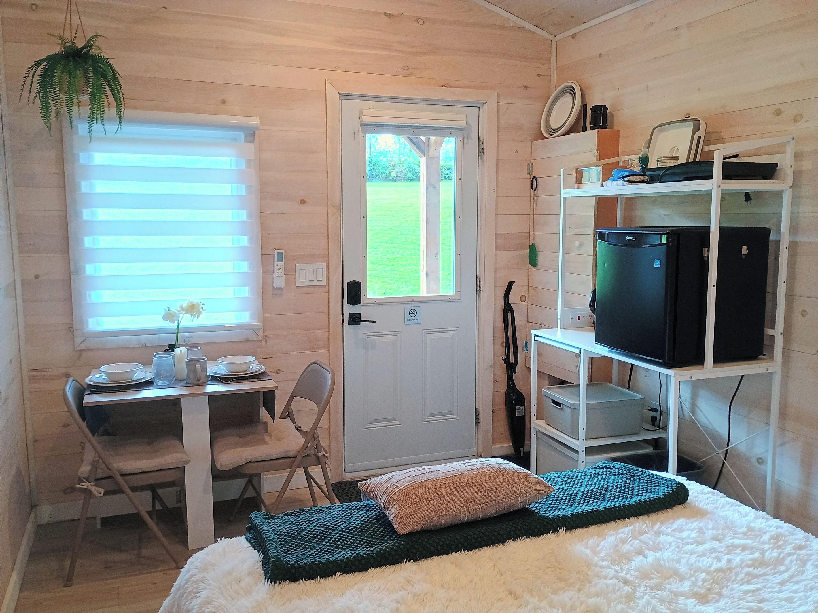 A compact room with a door, a table with chairs, a bed with pillows, a shelf with appliances, and a window with blinds.