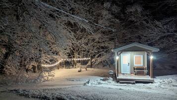 Una piccola baita con un portico, circondata da alberi innevati e una ghirlanda di luci.