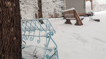 A metal bench with intricate designs, partially covered in snow, in a snowy outdoor setting with trees and a building in the background.