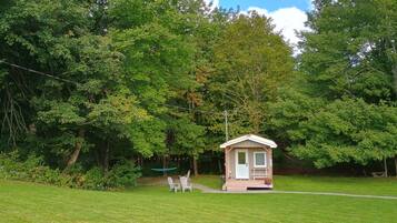 Une petite construction en bois, dotée d’une porte et de fenêtres, entourée d’une zone herbeuse et d’arbres.