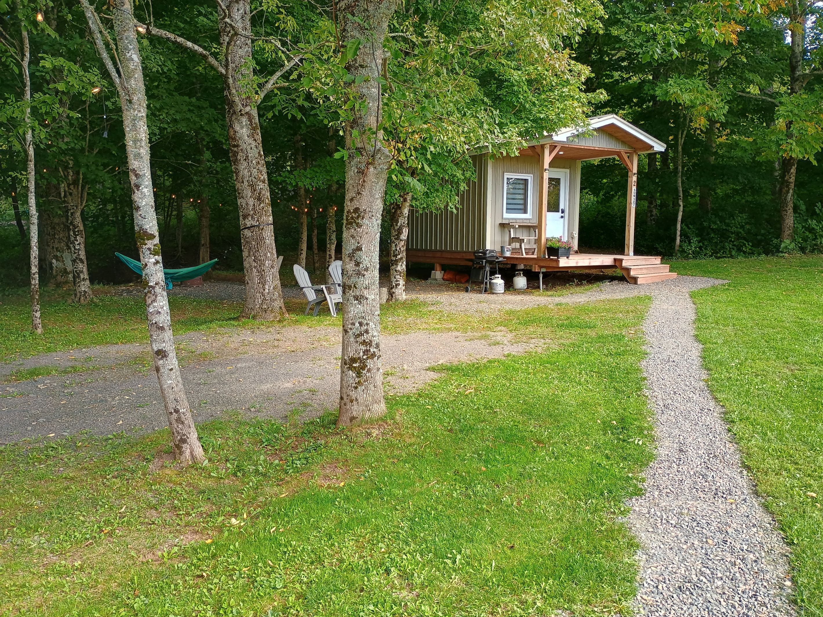 A small wooden cabin with a porch, surrounded by trees and a gravel path leading to it.