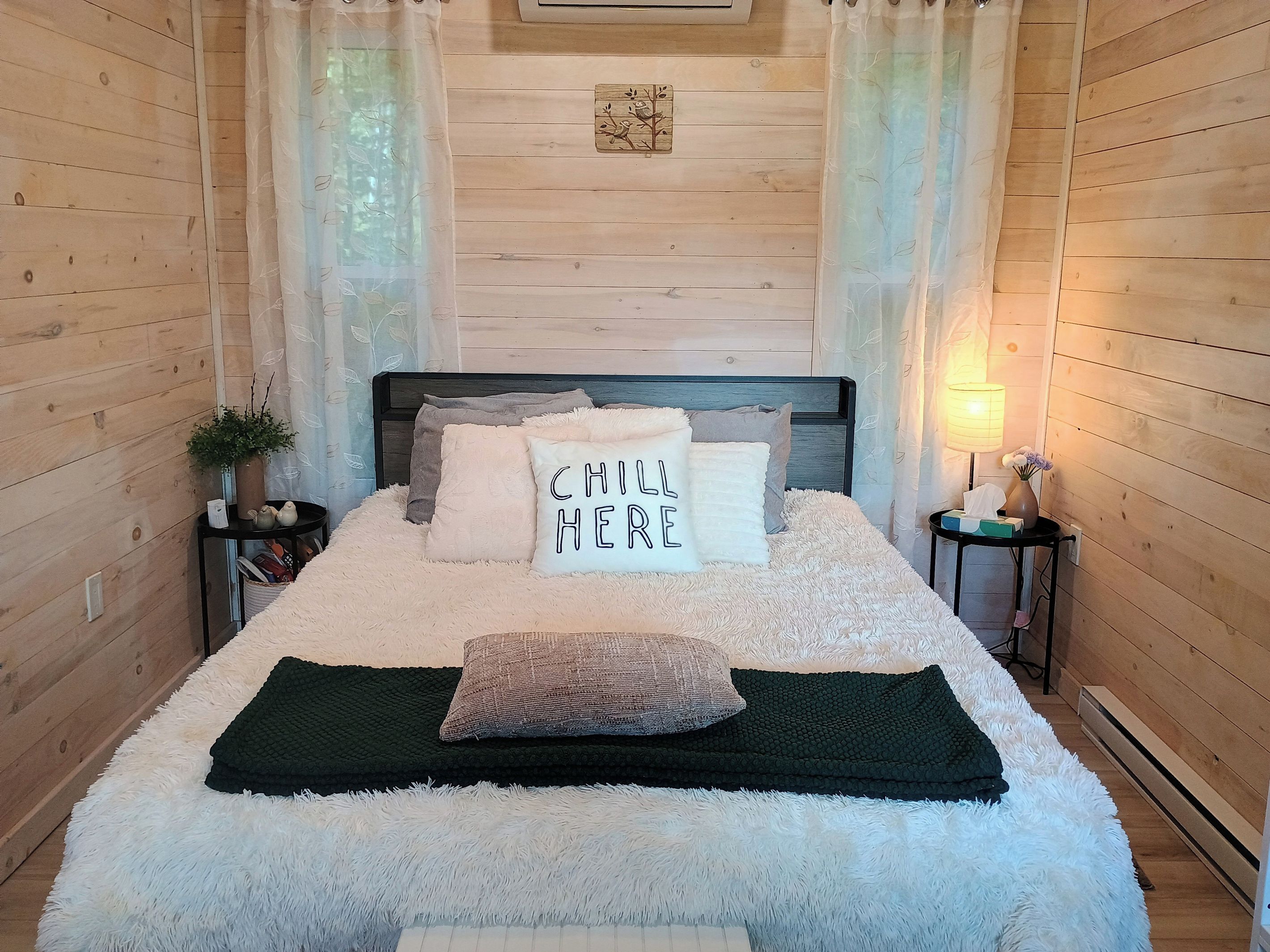 A cozy bedroom with a wooden headboard, a fluffy white bedspread, and a pillow with 'CHILL HERE' text.