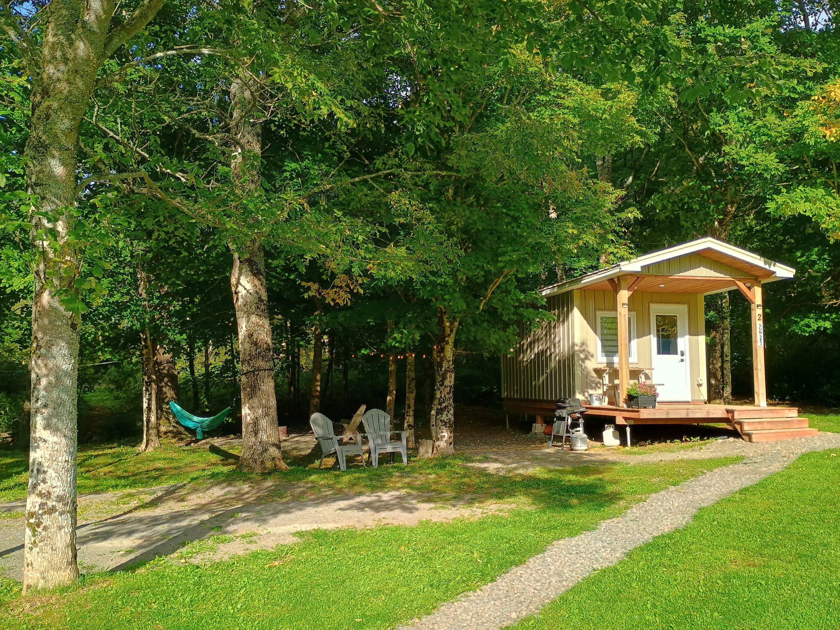 A small wooden cabin with a porch, surrounded by trees. There is a hammock and a picnic table with chairs.