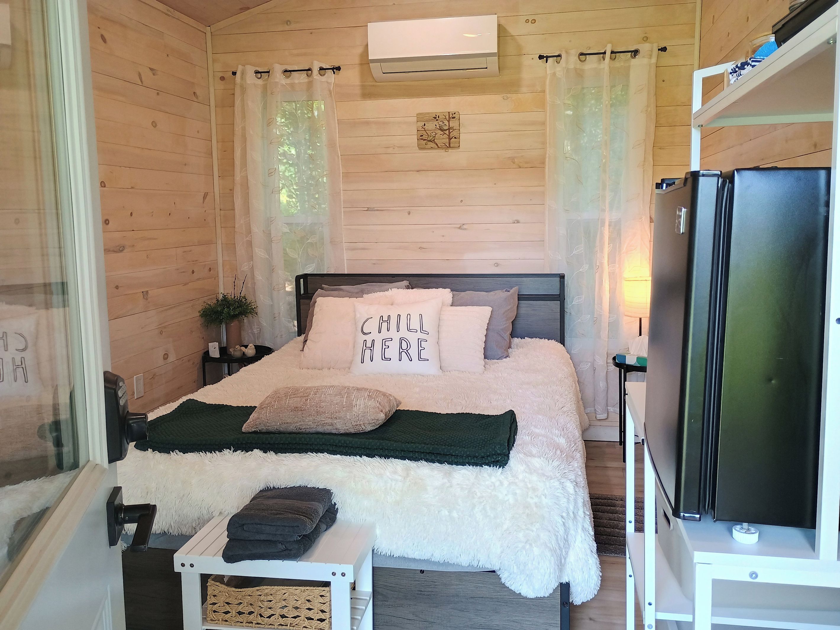 A cozy room with a bed, a small table, a refrigerator, and a window with curtains.