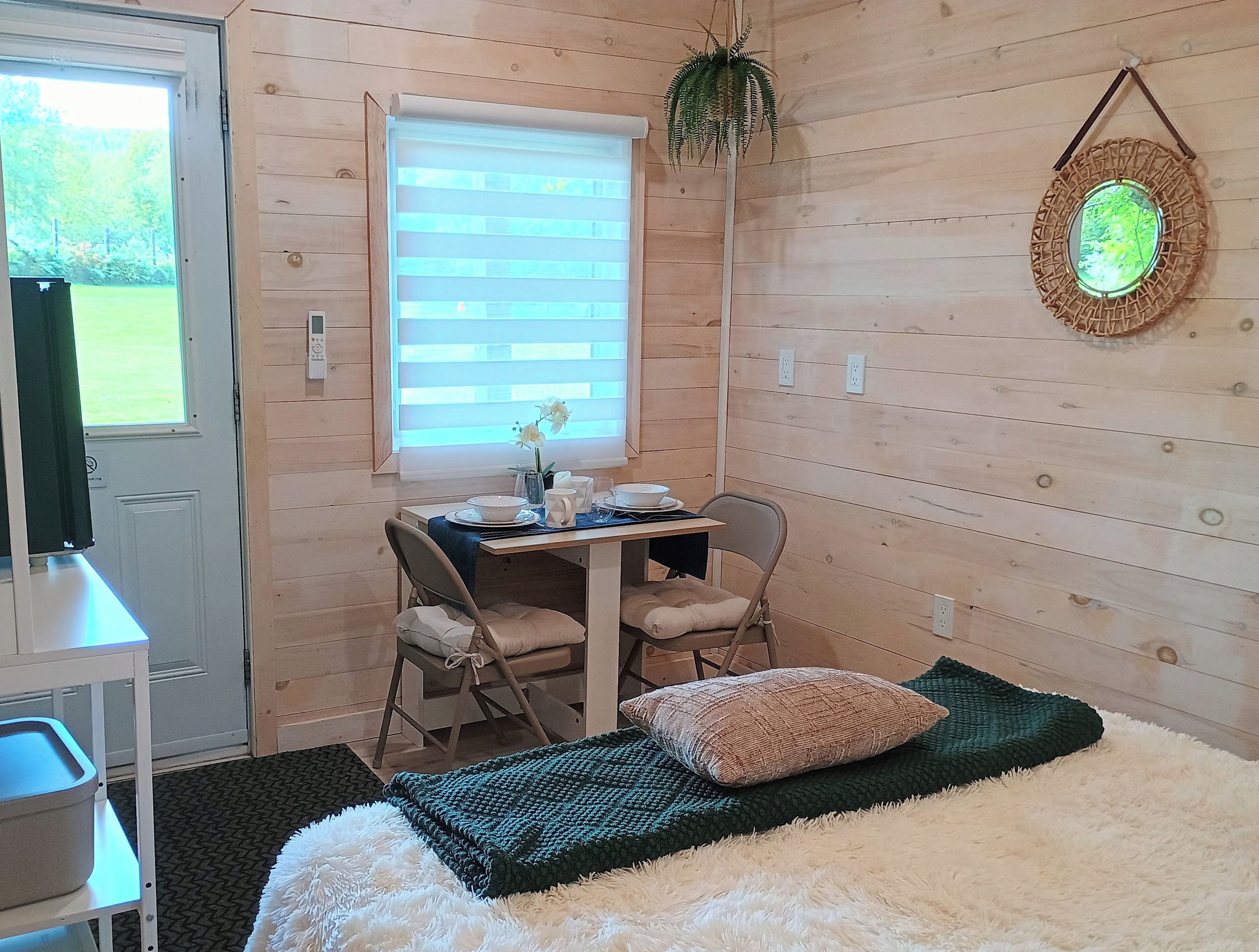 A room with a bed, a table with chairs, a window with blinds, and a hanging plant.
