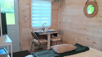 A room with a bed, a table with chairs, a window with blinds, and a hanging plant.