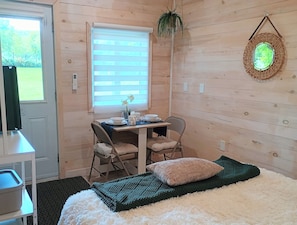 A room with a bed, a table with chairs, a window with blinds, and a hanging plant.