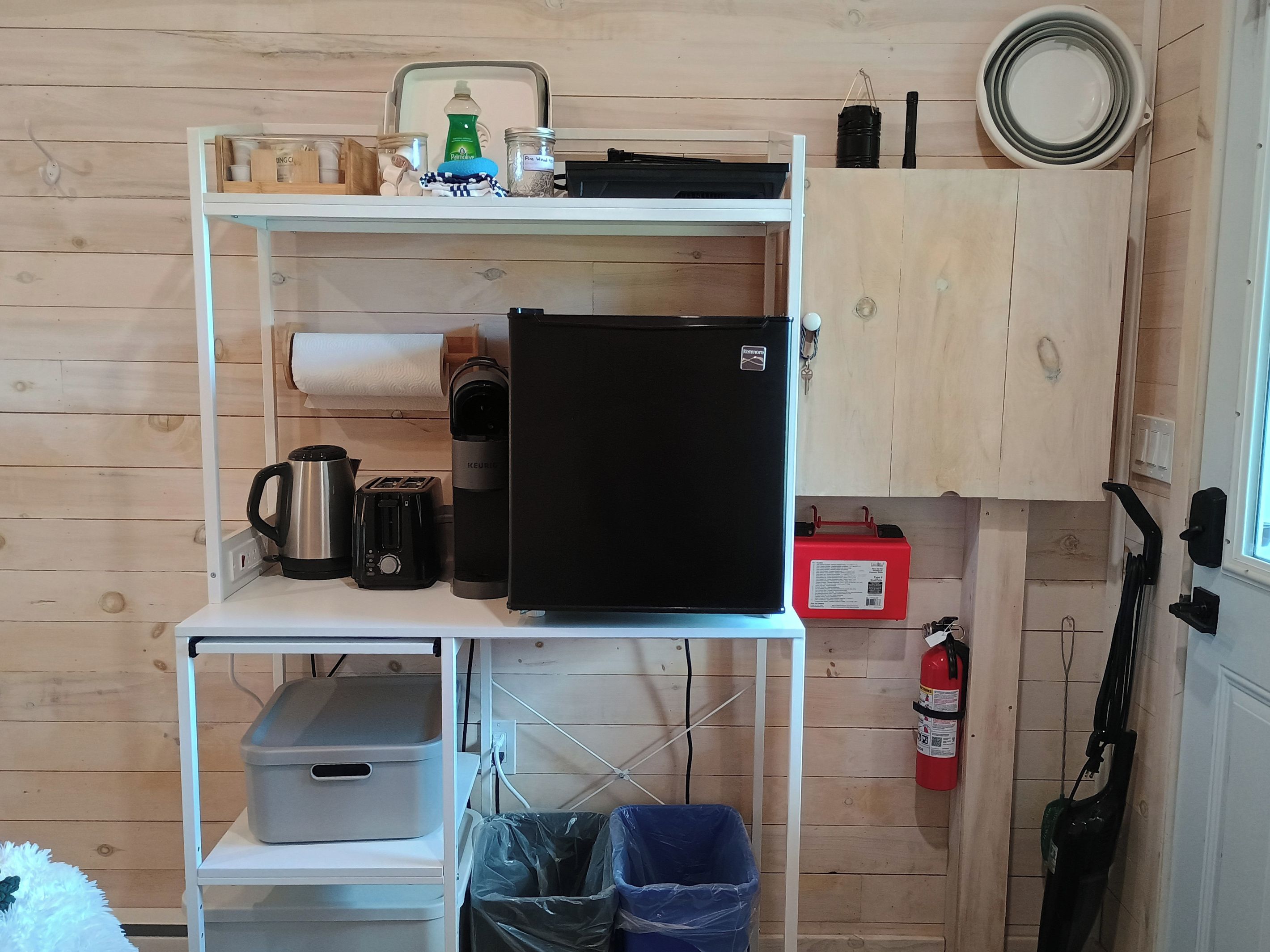A compact kitchen with a black refrigerator, a kettle, a toaster, and a coffee maker on a white shelf.